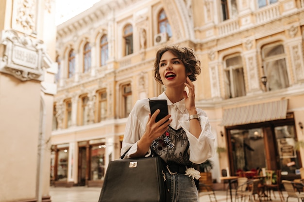 Femme à la mode en chemisier léger avec de la dentelle tenant un sac à main sombre et un téléphone en ville. Femme aux cheveux ondulés avec des lèvres brillantes détourne le regard sur la rue.