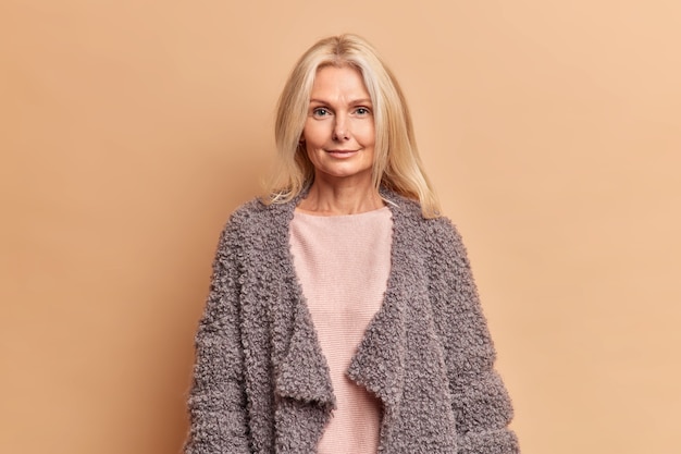 Femme à la mode de cinquante ans aux cheveux blonds vêtue d'un pull et d'un manteau chaud regarde directement à l'avant avec une expression sérieuse pose contre le mur beige reste belle à tout âge