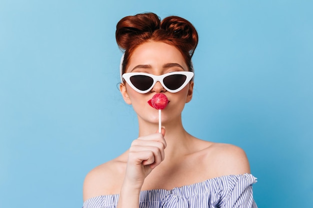 Femme à la mode dans des lunettes de soleil noires léchant la sucette Vue de face de pin-up dame avec des bonbons isolé sur fond bleu