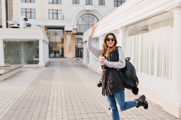 Femme à la mode drôle montrant de vraies émotions positives dans le centre-ville. Jeune femme avec du café à emporter, voyageant avec sac et appareil photo, portant un pull en laine, des lunettes de soleil, s'amusant. Place pour le texte.