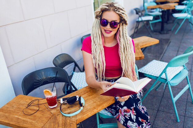 Femme à la mode élégante avec des dreadlocks blancs dans des lunettes de soleil tenant un cahier et passant son temps libre dans un restaurant moderne. Smoothie frais et écouteurs sur table.
