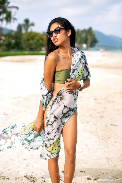 Femme à la mode élégante posind à la plage exotique tropicale, vacances de luxe, portant des lunettes de soleil en bikini et des vêtements de plage.