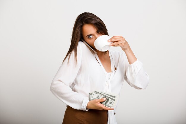 Femme de mode joyeuse, boire du café, garder de l'argent et parler au téléphone