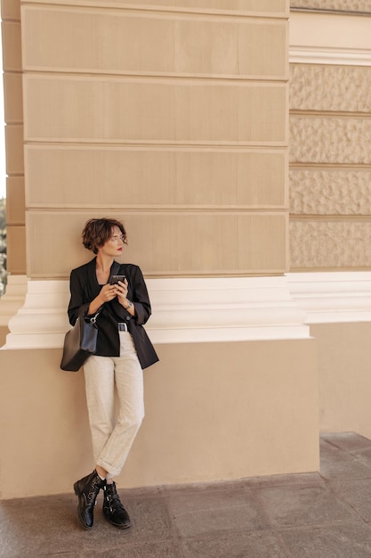 Femme à la mode en pantalon blanc, veste noire et bottes posant à l'extérieur