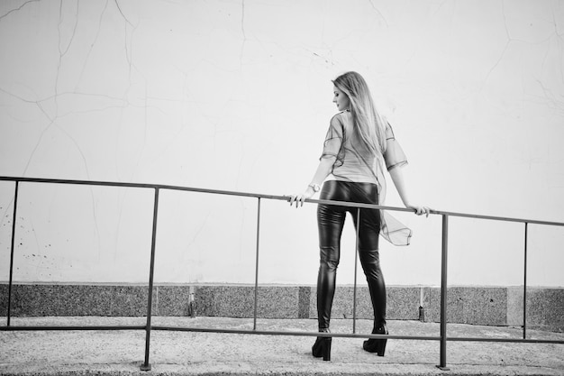 Femme à la mode regarde chemise blanche vêtements transparents noirs pantalons en cuir posant dans la rue contre les garde-corps en fer Concept de fille de mode