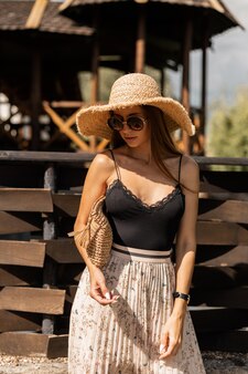 Femme modèle à la mode dans des lunettes de soleil rondes vintage avec un chapeau de paille avec un sac à main tissé dans une élégante lingerie en dentelle noire et une jupe se promène près de la plage pendant les vacances d'été