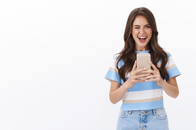 Femme moderne élégante et effrontée prenant un selfie dans un miroir, se faisant un clin d'œil ambitieuse et séduisante, souriante ravie, essayez un nouvel appareil photo pour smartphone, photographiant près d'un mur blanc