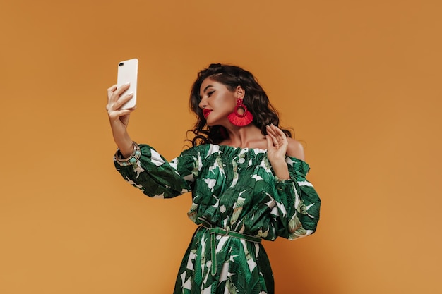 Femme moderne à la mode avec des cheveux bouclés et des boucles d'oreilles ren cool en robe d'été verte avec une ceinture fine fait selfie sur fond orange