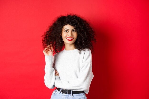 Femme moderne en sweat-shirt, jouant avec les cheveux bouclés et regardant de côté l'espace vide, debout sur fond rouge.