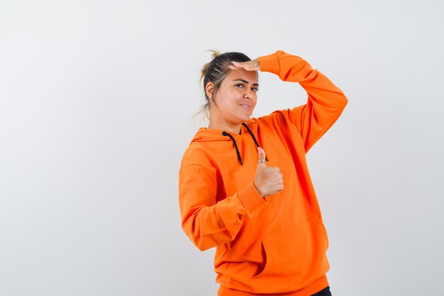 femme montrant le pouce vers le haut, regardant la caméra avec la main sur la tête en sweat à capuche orange et l'air mignon