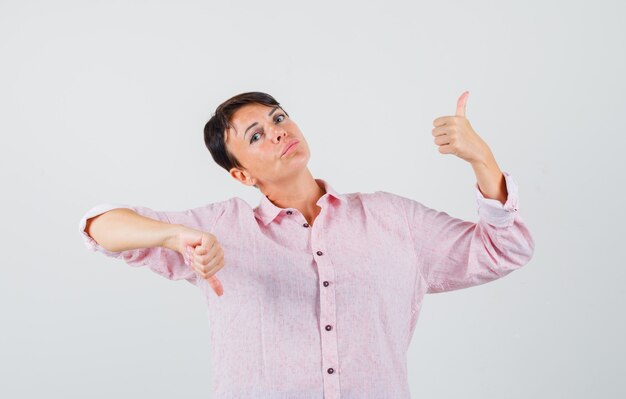 Femme montrant les pouces de haut en bas en chemise rose et à la vue de face, hésitante.