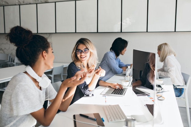 Photo gratuite femme mulâtre bouclée en t-shirt gris explique quelque chose à une amie blonde. portrait intérieur d'étudiants internationaux avec des ordinateurs portables se préparant pour le test ensemble.