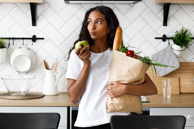 Une femme mulâtre réfléchie tient un paquet plein de légumes frais dans une main et une pomme mordue dans l'autre, sur la cuisine blanche moderne