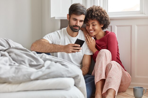 Femme multiethnique amicale, homme a lu du texte ou des nouvelles positives sur Internet sur téléphone mobile, vêtu de vêtements domestiques décontractés, assis sur le sol près du lit, connecté à Internet sans fil, temps de loisirs