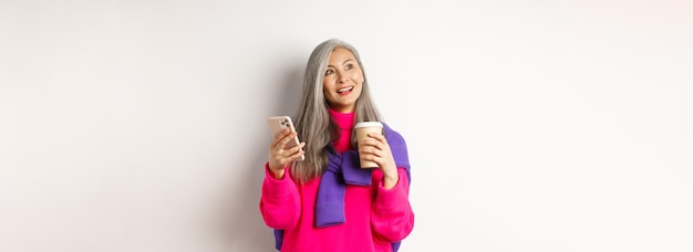 Femme mûre asiatique à la mode avec des cheveux gris buvant du café et utilisant un smartphone regardant en haut à gauche