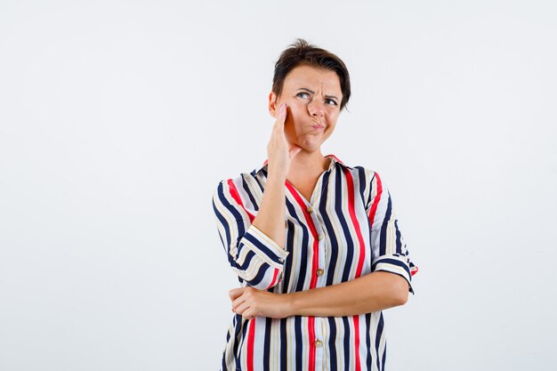 Femme mûre en chemisier rayé appuyé contre la joue sur la paume tout en pensant à quelque chose, tenant la main sur la taille et regardant pensif, vue de face.