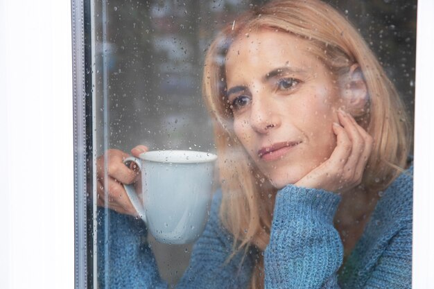 Femme mûre regardant par la fenêtre pendant qu'il pleut et buvant du thé