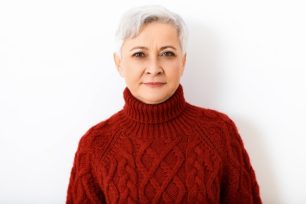 Femme mûre à la retraite aux cheveux gris sérieux isolés en pull à col roulé tricoté ayant une expression faciale concentrée ou suspecte stricte, posant contre un mur blanc vierge