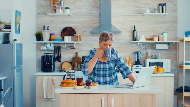 Femme mûre tapant sur un ordinateur portable dans la cuisine pendant le petit-déjeuner et buvant du café. Personne âgée à la retraite travaillant à domicile, télétravaillant à l'aide d'une communication en ligne sur Internet à distance sur une technologie moderne