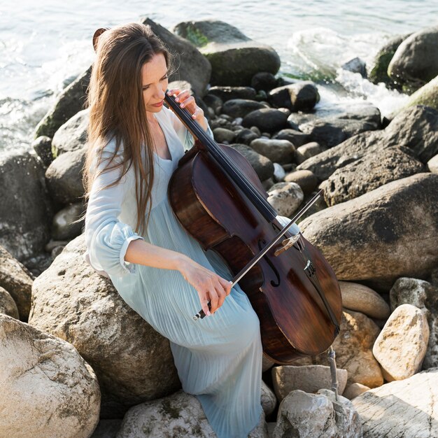 Femme musicienne sur des rochers à jouer du violoncelle au bord de la mer