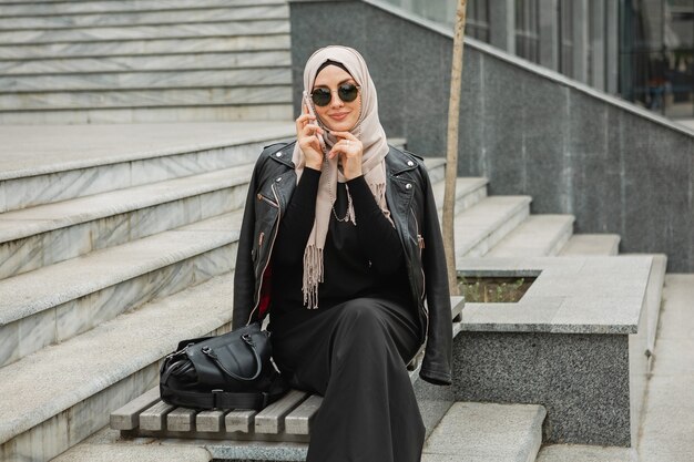 Femme musulmane élégante moderne en hijab, veste en cuir et abaya noire marchant dans la rue de la ville parlant sur smartphone