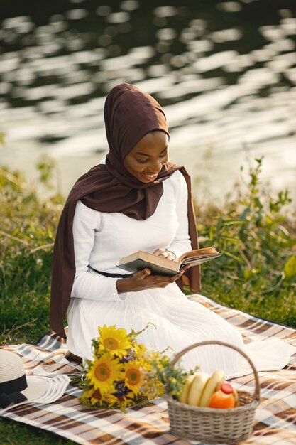 Une femme musulmane est assise sur la couverture de pique-nique à carreaux près de la rivière et lit un livre