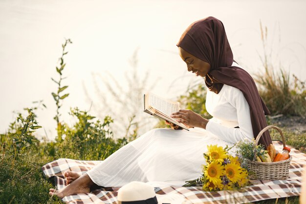 Une femme musulmane est assise sur la couverture de pique-nique à carreaux près de la rivière et lit un livre