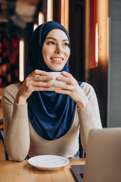 Femme musulmane moderne buvant du thé et travaillant sur ordinateur dans un café