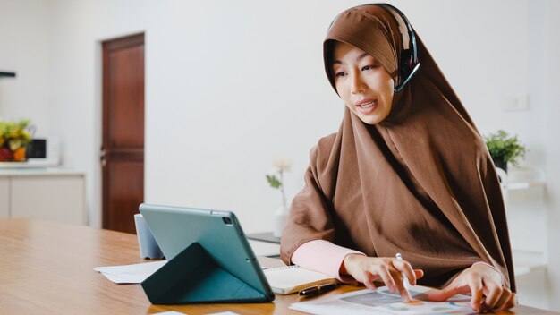 une femme musulmane porte un casque à l'aide d'une tablette numérique parle à ses collègues du rapport de vente lors d'une conférence vidéo tout en travaillant à domicile dans la cuisine.