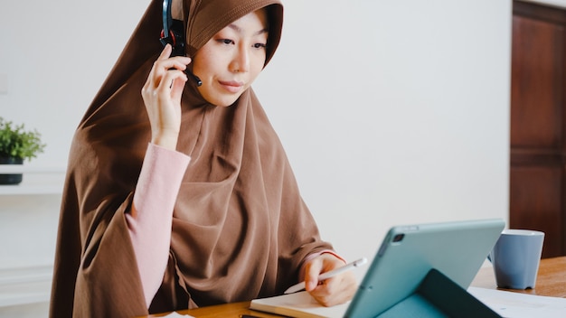 Une Femme Musulmane Porte Un Casque à L'aide D'une Tablette Numérique Parle à Ses Collègues Du Rapport De Vente Lors D'une Conférence Vidéo Tout En Travaillant à Domicile Dans La Cuisine.