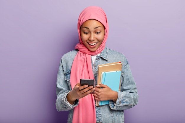 Une femme musulmane positive regarde un appareil smatphone, porte un hijab rose, une veste en jean