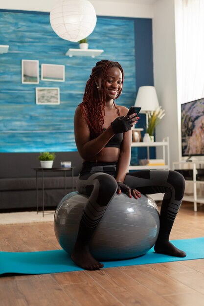 Femme noire athlétique sportive relaxante sur un ballon de stabilité vérifiant les médias sociaux à l'aide d'un smartphone après un exercice d'entraînement intense, dans le salon de la maison, assise sur un tapis de fitness pour la force musculaire.