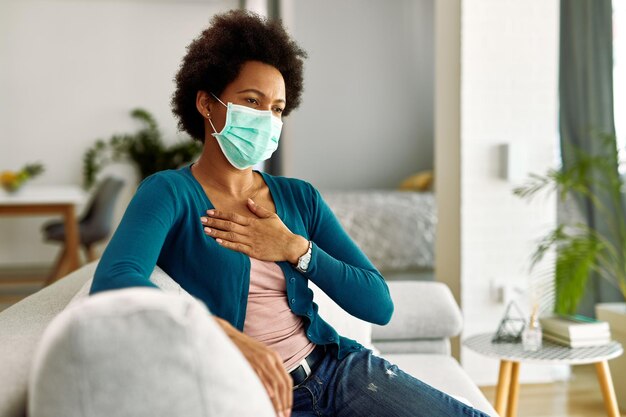 Femme noire portant un masque facial et tenant sa poitrine dans la douleur alors qu'elle était assise sur le canapé dans le salon