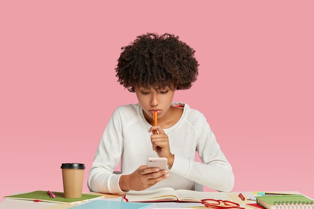 Une femme noire regarde sérieusement son téléphone intelligent, porte un pull blanc, garde un stylo en main
