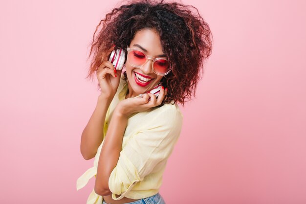 Femme noire timide à la peau brillante riant tout en se détendant avec de la bonne musique. Adorable fille mulâtre avec maquillage élégant relaxant dans les écouteurs.