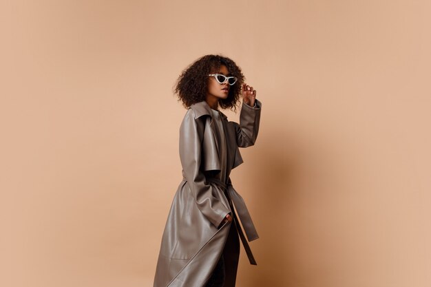 Femme noire en veste de cuir gris tendance posant sur fond beige en studio. Look mode hiver et automne.