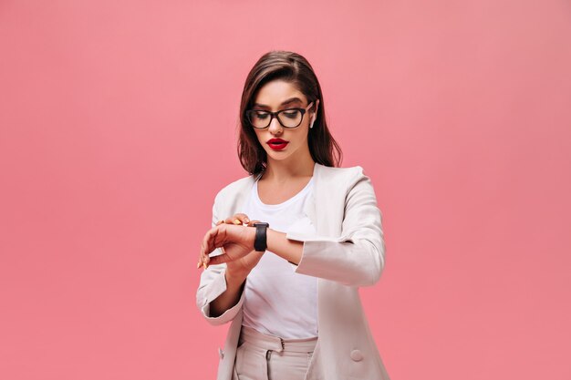 Femme occupée en veste beige regarde sa montre à main. Brunette avec des lèvres brillantes rouges dans des verres et des écouteurs blancs posant sur fond isolé.