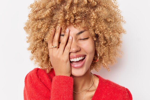 Une femme optimiste et heureuse aux cheveux bouclés couvre le visage avec la main rit joyeusement de quelque chose vêtu d'un pull rouge décontracté se sent fou de joie exprime des émotions positives isolées sur fond blanc.