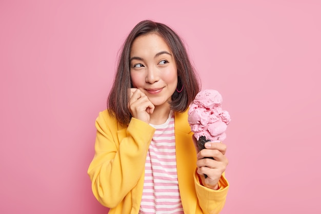 Une femme orientale rêveuse regarde agréablement loin avec une expression réfléchie tient une grande glace savoureuse pense à quelque chose qui garde la main sur le visage vêtue de vêtements élégants isolés sur un mur rose