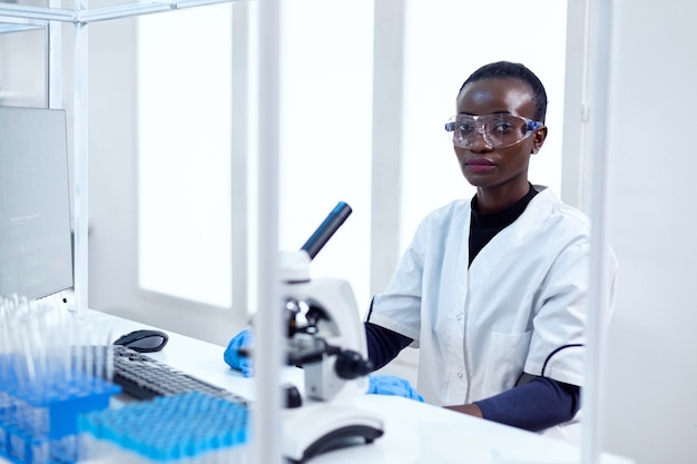 Femme d'origine africaine travaillant dans un établissement de santé assise sur son lieu de travail avec des lunettes de protection. scientifique noir de la santé dans un laboratoire de biochimie portant un équipement stérile.