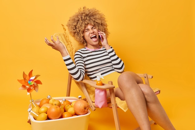 Photo gratuite une femme outragée crie avec colère fait un appel téléphonique porte un pull rayé et un short a une conversation ennuyeuse tout en se reposant à la plage sur une chaise longue utilise un réfrigérateur portable sur fond jaune