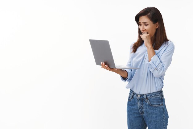 Femme paniquée mordant le poing debout nerveusement tenir l'écran fixe de l'ordinateur portable avec anxiété avoir un virus informatique supprimant un document de travail important posant un fond blanc inquiet et en détresse