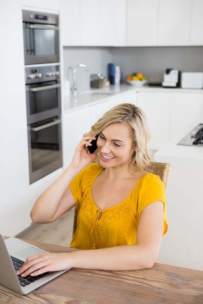 Femme parlant au téléphone mobile en utilisant un ordinateur portable dans la cuisine