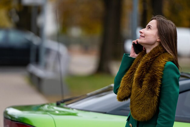 Une femme parlant au téléphone en se tenant près d'une voiture verte garée dans une rue de la ville en arrière-plan