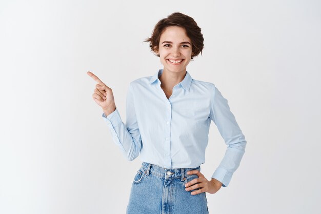 Femme pdg professionnelle souriante en chemise à col bleu, pointant le doigt vers la gauche et l'air confiant, debout sur un mur blanc