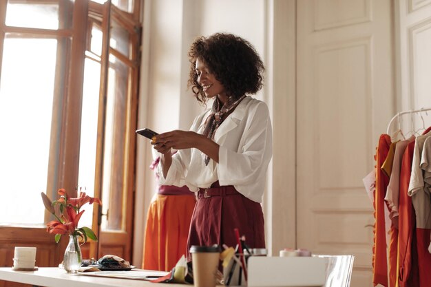 Une femme à la peau foncée bouclée à la mode en pantalon bordeaux et chemisier blanc prend une photo d'échantillons de textile tient un téléphone et sourit au bureau