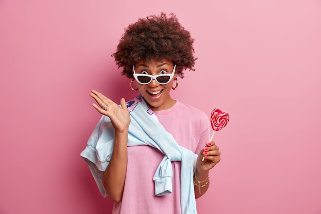 Photo gratuite une femme à la peau foncée positive a les cheveux bouclés, tient une sucette, s'amuse avec des amis pendant la journée, porte des lunettes de soleil, aime les bonbons, pose