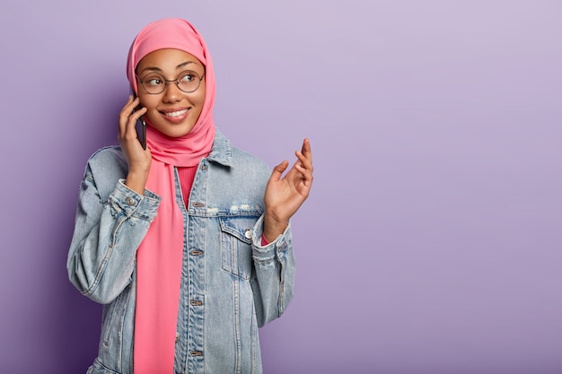 Photo gratuite une femme à la peau foncée positive porte un foulard musulman traditionnel sur la tête, garde le téléphone portable près de l'oreille, profite d'une conversation agréable, fait des gestes tout en disant quelque chose d'excitant à l'interlocuteur isolé sur violet