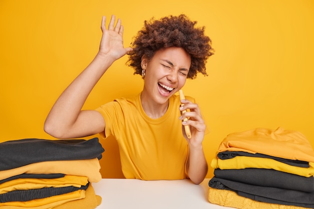 Une femme à la peau foncée ravie lève la paume chante une chanson tient un smartphone comme si le microphone était assis à table avec du linge plié isolé sur un mur jaune. Femme occupée plie les vêtements après le lavage