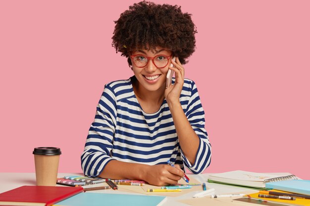 Une femme à la peau foncée réussie avec une coupe de cheveux afro, vêtue de vêtements à rayures, a une conversation téléphonique agréable tout en dessine quelque chose dans le bloc-notes, boit du café à emporter, se sent satisfaite et inspirée